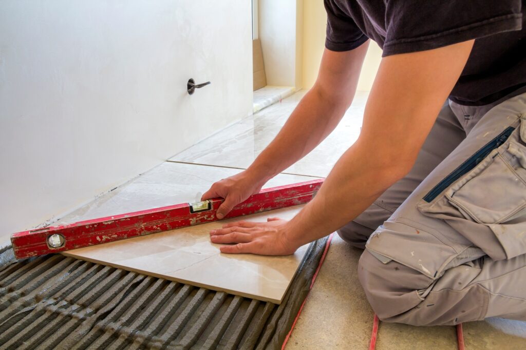 Young worker tiler installing ceramic tiles using lever on cement floor with heating